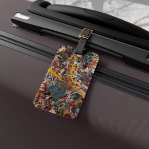 May include: A rectangular luggage tag featuring a vibrant paint splatter pattern in shades of yellow, teal, red, and white. The tag is attached to a dark gray suitcase handle by a black strap with a gold-toned metal buckle.