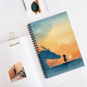 Può includere: Quaderno a spirale con un'illustrazione ad acquerello di un drago blu di fronte a una piccola figura contro un tramonto. Il quaderno ha una rilegatura a spirale nera ed è aperto su una superficie bianca.