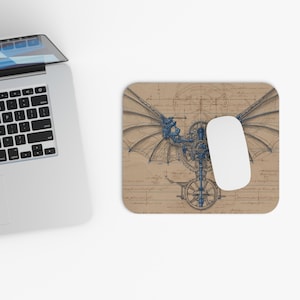 May include: A tan mousepad featuring a detailed blue and brown steampunk-style mechanical winged creature design. A white computer mouse rests on the pad, next to a laptop keyboard.