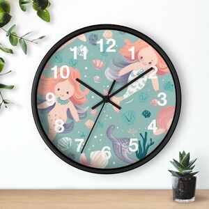 May include: A round wall clock with a black frame and a teal background featuring a pattern of mermaids, seashells, and coral. The clock face displays large white numbers from 1 to 12 and has three black hands to indicate the time.