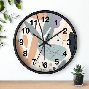 Könnte beinhalten: Runde Wanduhr mit schwarzem Rahmen. Das Zifferblatt zeigt ein abstraktes Design in Pastellfarben, darunter Pfirsich, Blau und Lavendel, mit schwarzen Ziffern und Zeigern. Die Uhr befindet sich auf einer Holzoberfläche.