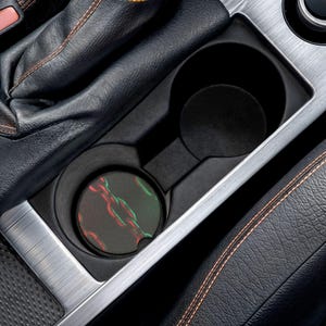 May include: A black car cup holder with a coaster featuring a chain design in red and green. The coaster is placed in the bottom of the cup holder. The car interior is black with silver trim and orange stitching.