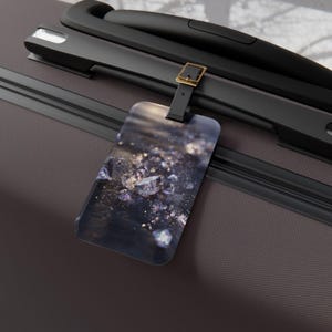 以下が含まれることがあります： 紫と金のきらめく結晶のような形が散りばめられた、暗い抽象的なプリントの長方形のラゲッジタグ。タグは、金色の金属製バックルが付いた黒いストラップで、ダークブラウンのスーツケースのハンドルに取り付けられています。