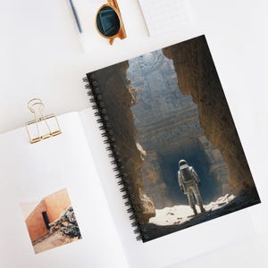 May include: Spiral-bound notebook with an illustration of an astronaut in a white spacesuit exploring a stone structure. The astronaut is standing in a large, open space with light shining from above. The notebook is open on a white surface.
