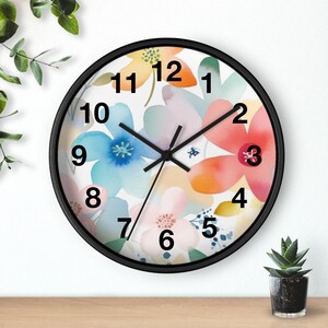 Puede incluir: Un reloj de pared redondo con marco negro y esfera blanca que presenta un patrón floral de acuarela en tonos azules, rosas, naranjas y amarillos. El reloj tiene manecillas negras y números negros del uno al doce en la esfera.