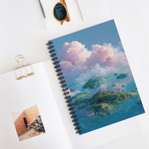 May include: Spiral-bound notebook featuring a dreamy landscape with fluffy pink clouds, blue water, and floating islands. The notebook has a black spiral binding and a colorful cover design. A gold paper clip and sunglasses are also visible.