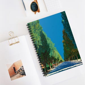 May include: Spiral-bound notebook featuring a vibrant landscape painting of tall green trees lining a road under a bright blue sky. The notebook is open, revealing the artwork and a gold paper clip.