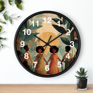 Pode incluir: Relógio de parede redondo com moldura preta, exibindo uma ilustração de figuras de mãos dadas contra um fundo de vegetação e um sol poente. Os números brancos marcam as horas. Uma pequena planta em vaso está abaixo.