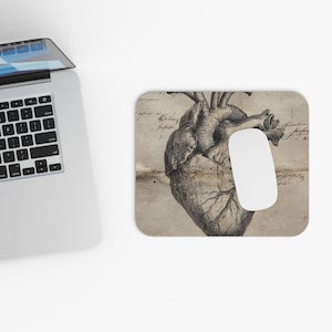 Op de afbeelding: Een computermuis en muismat met een gedetailleerde anatomische illustratie van een menselijk hart. De muismat heeft een vintage, verouderde papieruitstraling met handschrift. De muis is wit.