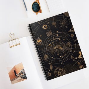 May include: A spiral-bound notebook with a black cover featuring a gold astrological design. The design includes zodiac symbols, constellations, and celestial elements. The notebook is open on a white surface, with a gold paper clip and sunglasses nearby.