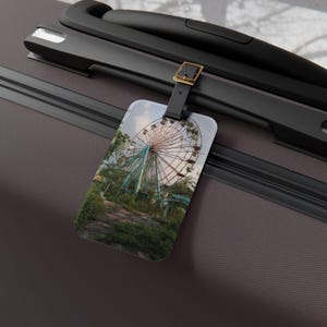 May include: A rectangular luggage tag featuring a printed image of a large, weathered Ferris wheel surrounded by overgrown greenery and trees. The tag is attached to a dark brown suitcase handle by a black strap with a gold-toned metal buckle.