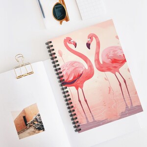 May include: Spiral-bound notebook featuring a vibrant illustration of two pink flamingos with long necks and legs. The notebook has a pink and peach background with a black spiral binding. Other items include a gold binder clip and sunglasses.