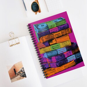 May include: Spiral-bound notebook with a vibrant cover featuring stacks of colorful, stylized dollar bills. The bills are in shades of blue, green, orange, and purple, with text on the bands. The notebook is on a white surface with a gold paper clip and sunglasses.