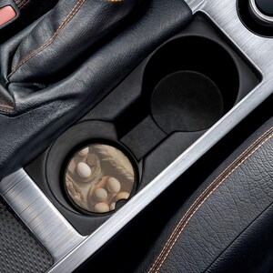 May include: A car cup holder with a coaster featuring a still-life image of eggs and wheat. The coaster is round and fits snugly in the cup holder. The car's interior is black with silver and orange accents.