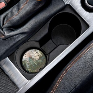 May include: A circular car cup holder coaster featuring a printed image of a Ferris wheel against a sky background. The coaster is placed inside a black plastic vehicle center console cup holder next to a black leather gear shift boot with stitching.