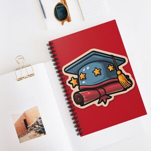 May include: A red spiral-bound notebook with a graduation cap and diploma sticker. The cap is blue with gold stars, and the diploma is red. The notebook is open on a white surface with a gold paper clip and sunglasses.