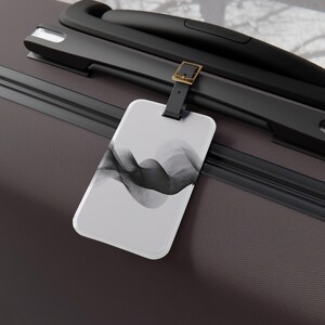 May include: A rectangular luggage tag featuring a black and white abstract design. It's attached to a black leather strap with a gold-coloured buckle. The tag rests on a dark brown suitcase.