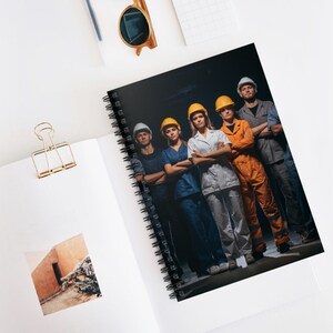May include: A spiral-bound notebook featuring a group of people in work attire, including hard hats and coveralls. The individuals are standing with their arms crossed, suggesting a team or workforce. The background is dark, emphasizing the subjects.