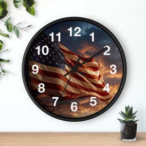 May include: Round black wall clock featuring the American flag waving against a sunset sky. The clock face has white numerals and black hands. A small potted plant sits on a wooden surface.