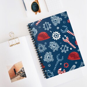 May include: Spiral-bound notebook with a navy blue cover featuring a pattern of gears, wrenches, and red hard hats. The wrenches have a red and white striped design. The notebook is open on a white surface.
