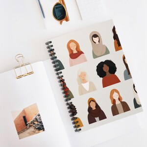 Puede incluir: Cuaderno de espiral con ilustraciones de rostros de mujeres diversas. La portada presenta una variedad de tonos de piel, peinados y vestimenta. El cuaderno está abierto, revelando una página en blanco y un clip dorado. Gafas de sol y un bolígrafo son visibles.