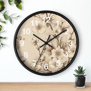 Op de afbeelding: Een ronde wandklok met een zwarte rand en een beige wijzerplaat met een gedetailleerde botanische illustratie van kersenbloesems in bruine inkt. De klok heeft grote witte cijfers van 1 tot 12 en dunne zwarte wijzers.