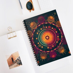 May include: Spiral-bound notebook with a colourful mandala design. The cover displays an intricate pattern with orange, purple, and gold details on a dark teal backdrop. The notebook is open, showing blank pages and a photograph.