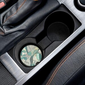 Könnte beinhalten: Ein runder Auto-Untersetzer mit einem abstrakten Design in Türkis, Weiß und Beige befindet sich in einem Getränkehalter im Auto. Der Untersetzer hat ein Muster aus organischen Formen und Linien. Der Innenraum des Autos ist schwarz mit silbernen und orangefarbenen Akzenten.