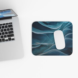 May include: A rectangular mousepad with a blue and white abstract design, featuring a white computer mouse. The mousepad has a textured, flowing pattern. A laptop is partially visible in the upper left corner.