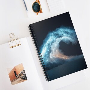May include: Spiral-bound notebook featuring a vibrant blue and white wave design against a dark background. The notebook is open, revealing a blank page and a gold binder clip. Sunglasses rest on the white surface.