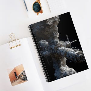 May include: Spiral-bound notebook featuring a detailed illustration of a futuristic space station. The artwork depicts a complex structure with metallic and weathered textures, set against a dark, starry background. The notebook is open on a white surface.