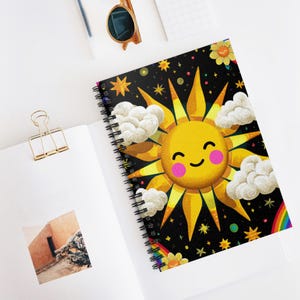 Puede incluir: Cuaderno de espiral con un diseño de sol sonriente, nubes, estrellas y un arcoíris sobre un fondo negro. El cuaderno está abierto sobre una superficie blanca, con un clip dorado y gafas de sol cerca. El cuaderno mide aproximadamente 20 cm de alto.