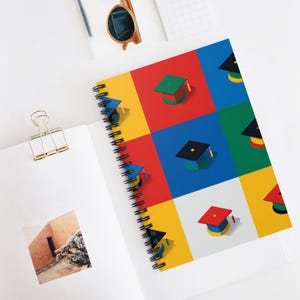 May include: A spiral-bound notebook with a colorful graduation cap design. The cover features a grid of red, yellow, blue, and green squares, each with a different colored graduation cap. The notebook is open, revealing blank pages.
