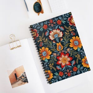 May include: A spiral-bound notebook with a dark blue cover featuring a vibrant floral pattern in orange, red, and blue. The notebook is open on a white surface, with a gold paper clip and sunglasses nearby. The design is reminiscent of vintage botanical illustrations.