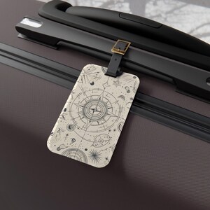 May include: A rectangular luggage tag with a cream-coloured background featuring black line art of a nautical compass, celestial stars, and abstract cosmic shapes. The tag is attached to a dark brown suitcase handle with a black strap and gold buckle.