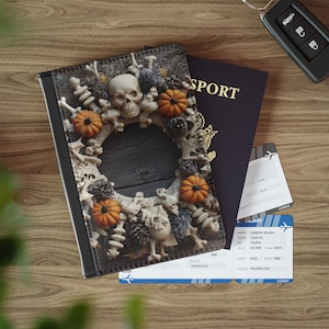 May include: A black passport cover with a decorative skull and pumpkin design. A dark blue passport, two plane tickets, and a car key are next to the cover on a wooden surface. The passport cover has a skull, pumpkins, and bones.