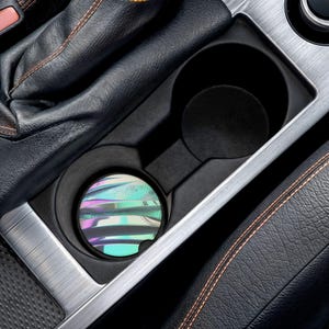 May include: A car cup holder with a colorful, iridescent coaster. The coaster features a design of horizontal lines in shades of purple, blue, and green. The cup holder is black and sits within a silver and black car interior.