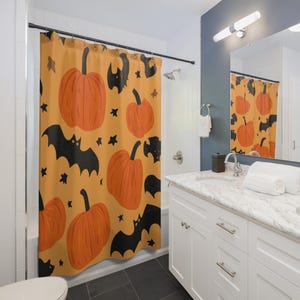 Op de afbeelding: Halloween douchegordijn met een patroon van oranje pompoenen, zwarte vleermuizen en sterren op een lichtoranje achtergrond. Het gordijn hangt in een badkamer met witte kasten en een marmeren aanrecht.