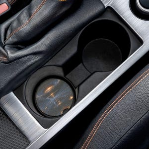 May include: A circular car cup holder coaster featuring a dark space-themed print with glowing golden light patterns. The coaster is placed inside a black plastic dual cup holder console, which is surrounded by brushed metal trim and black leather.