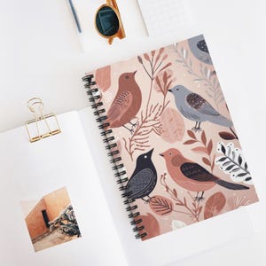 May include: A spiral-bound notebook featuring a bird and botanical design. The cover showcases illustrations of birds in shades of brown, gray, and black, set against a light pink background with leaves and branches. The notebook is open on a white surface.