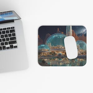 May include: A rectangular mousepad featuring a futuristic city scene with domed structures and a white computer mouse. The background shows a night sky with mountains and a body of water. The mousepad is next to a laptop.