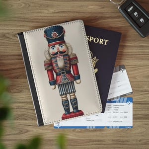 Puede incluir: Una funda para pasaporte con una ilustración de un soldado cascanueces en rojo, azul y blanco. Un pasaporte azul oscuro, billetes de avión y una llave de coche sobre una superficie de madera. Accesorios de viaje.