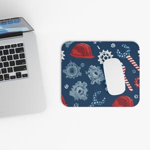 May include: A rectangular mousepad with a navy blue background featuring a pattern of red hard hats, gray gears, and wrenches with red and white stripes. A white computer mouse sits on the pad near a laptop.