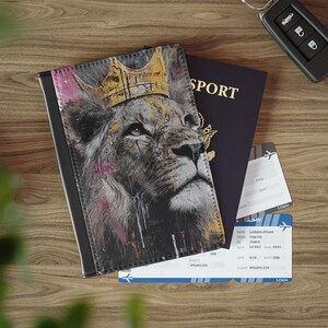 May include: Black passport holder featuring a lion wearing a gold crown. Beside it are a dark blue passport, plane tickets, and a car key. The lion graphic has pink and white paint splatters.