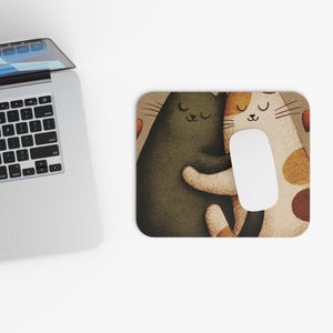 May include: A rectangular mousepad featuring a whimsical illustration of two hugging cats. One cat is dark green, the other is white with brown spots. A white computer mouse rests on the pad. The background is a light brown color.