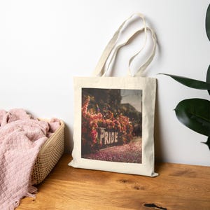 Peut inclure: Un sac fourre-tout beige avec une image rectangulaire affichant le mot "PRIDE" sur une enseigne entourée de fleurs roses. Le sac est posé sur une surface en bois, avec un panier de tissu rose à gauche.