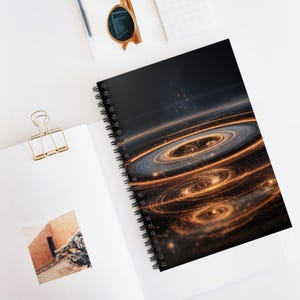 May include: A spiral-bound notebook with a black cover showing a cosmic design of swirling gold and silver rings against a dark backdrop. The notebook is open on a white surface, with a gold paper clip and sunglasses nearby.