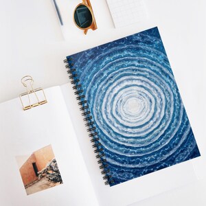 May include: Spiral-bound notebook featuring a blue and white watercolor design. The cover art depicts concentric circles resembling water ripples. The notebook is open, revealing blank pages, and rests on a white surface with a gold paper clip.