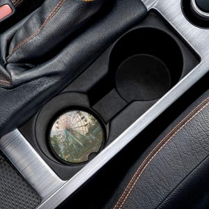 May include: A circular car cup holder coaster featuring a printed image of a Ferris wheel against a sky background. The coaster is placed inside a black plastic vehicle center console cup holder next to a black leather gear shift boot with stitching.