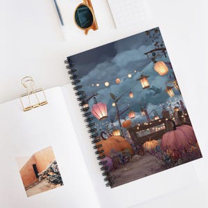 May include: A spiral-bound notebook featuring a colorful illustration of a nighttime pumpkin patch scene. The artwork includes glowing lanterns, pumpkins, and a cloudy sky. The notebook is open on a white surface with a gold paper clip.